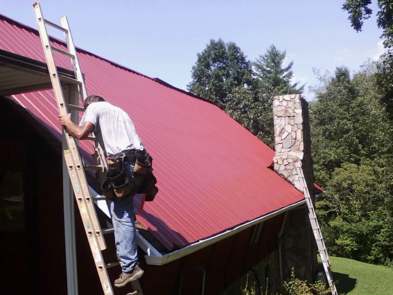 Roofer on a ladder beside a red metal roof and stone chimney in Oak Ridge